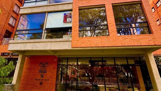 A multi-story building with a brick facade, large windows, and a ground-level entrance with a sign. - 605-Estudio luminoso con balcón  (Bogotá)