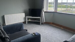Ein Wohnzimmer mit grauem Sofa, einer weißen TV-Konsole und einem Fenster mit Blick auf Grünflächen und eine Steinmauer.