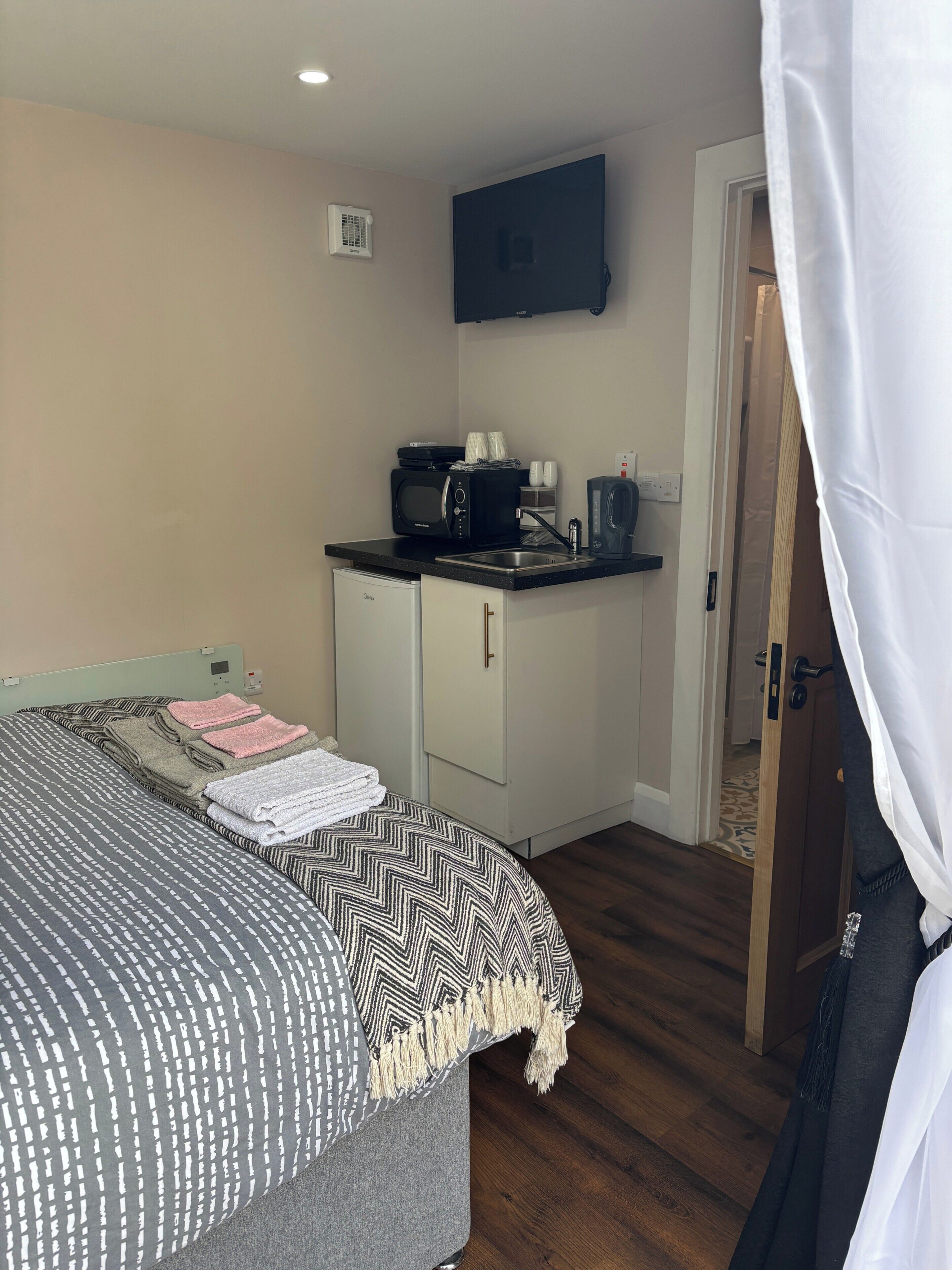 A compact hotel room with a bed, a kitchenette, a flat-screen TV, and a door leading to another room.