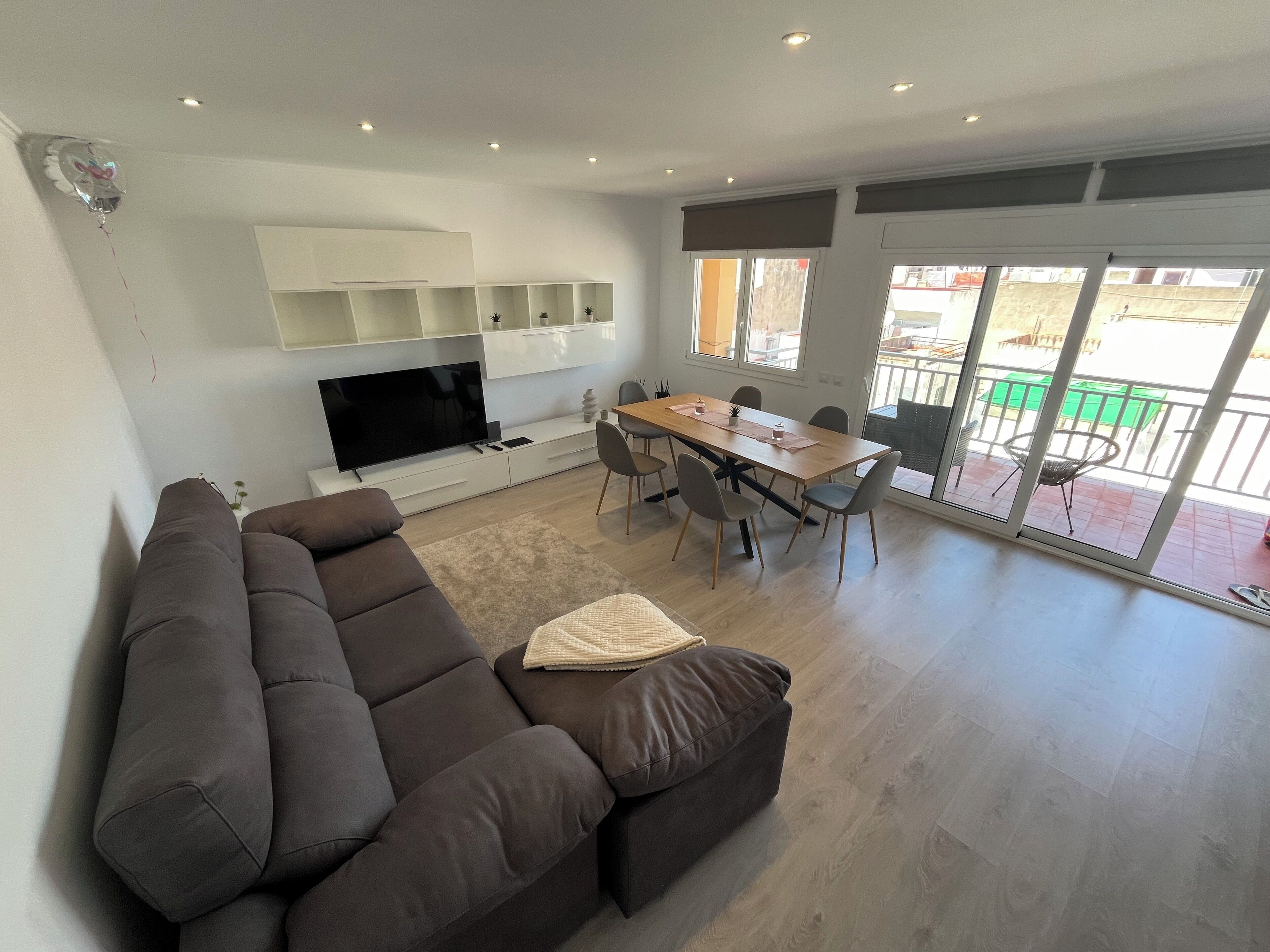 A modern living room with a large dark brown sofa, a flat-screen TV, a wooden dining table with chairs, and a balcony with a green table and chairs.