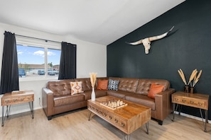 A living room with a brown leather sofa, a wooden coffee table, and a large window with curtains.