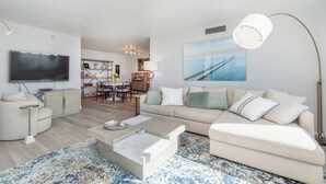 A modern living room with a sofa, coffee table, and TV. There is a dining area in the background. - Beachfront Bliss: 2-Bed/2-Bath Condo with Stunning Views! (Fort Myers Beach)