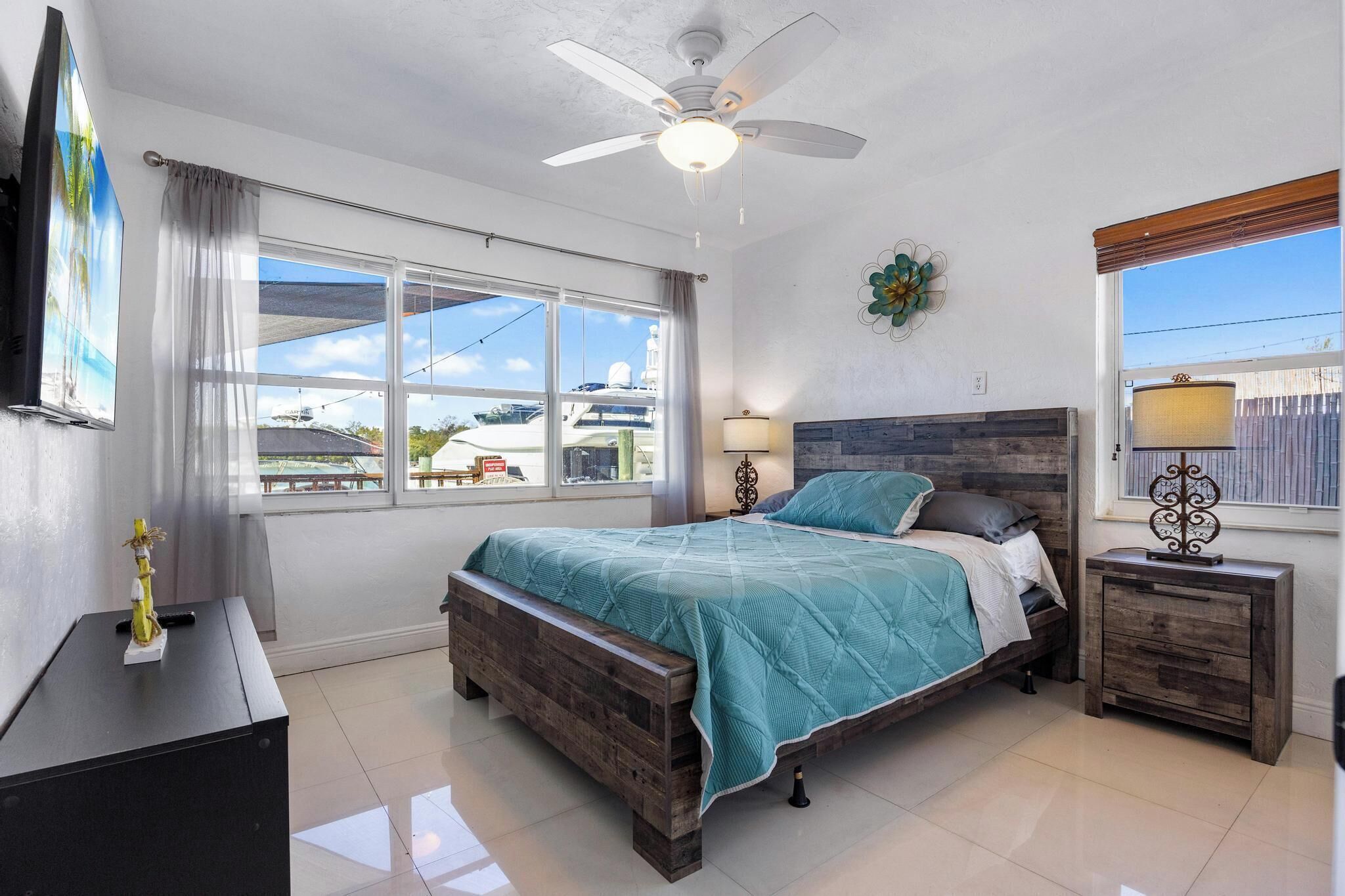 A bedroom with a bed, a nightstand, a ceiling fan, a window, and a wall-mounted TV.