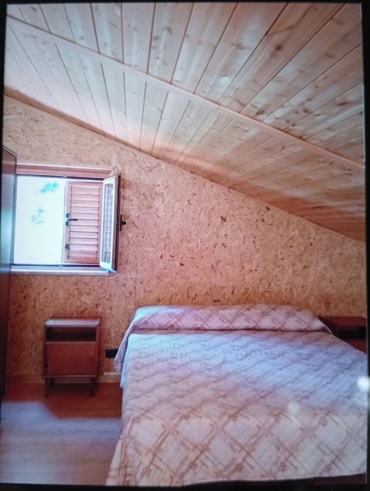 A bedroom with a sloped ceiling, a window with shutters, a bed with patterned bedding, and a small wooden nightstand.