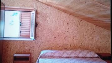 A bedroom with a sloped ceiling, a window with shutters, a bed with patterned bedding, and a small wooden nightstand.