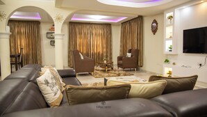 A living room with a brown leather sofa, two armchairs, a coffee table, and a TV mounted on the wall. - Unique Green Lodge (Accra)