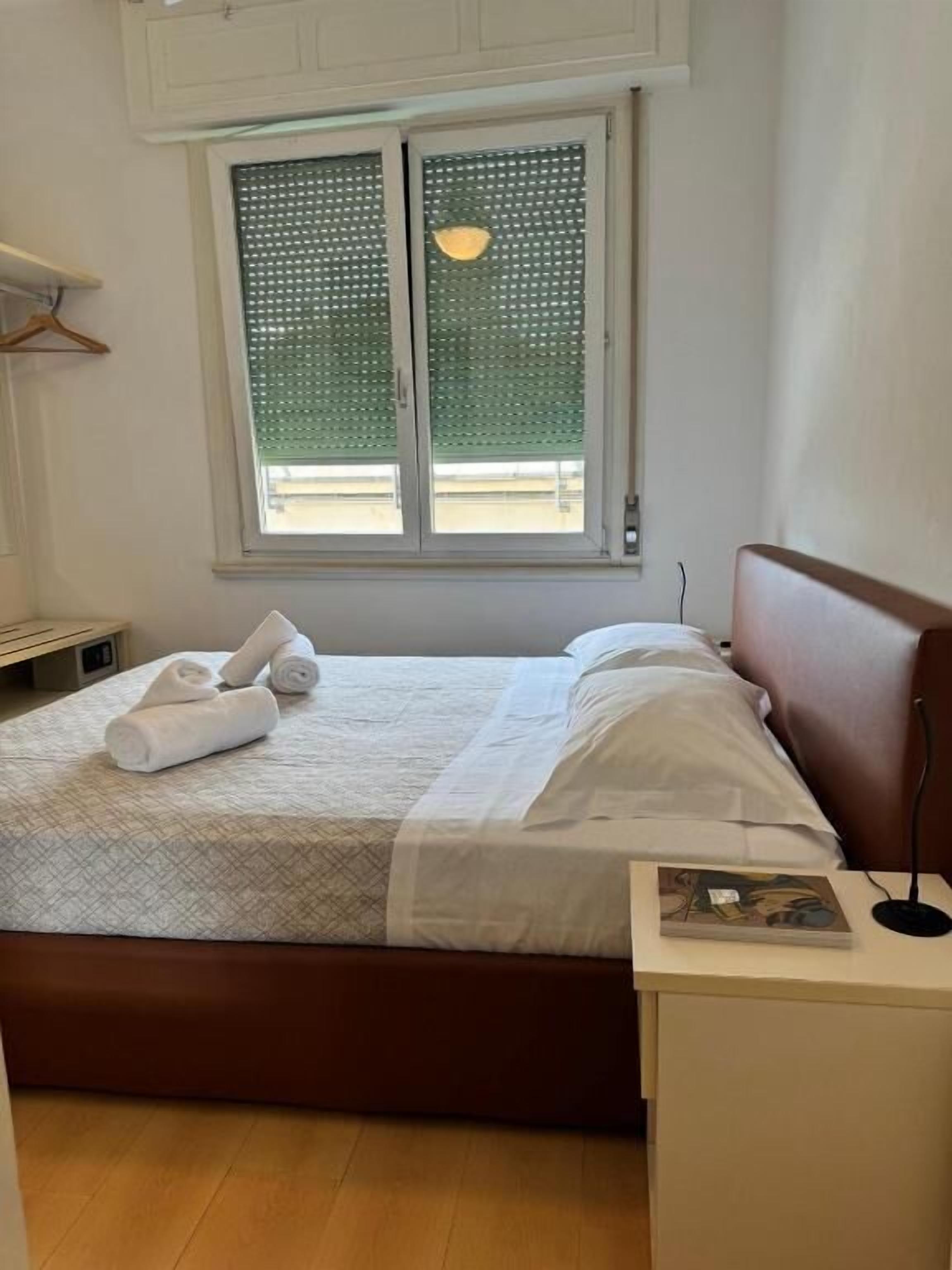 A neatly made bed with white linens and rolled towels, a bedside table with a lamp, and a window with blinds.