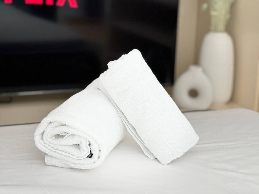 A neatly folded white towel on a bed with a television displaying the word "FLIX" in the background.
