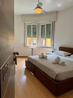Egy hálószoba, amelyben van egy ágy, egy mennyezeti ventilátor, egy rolós ablak és egy kis asztal.