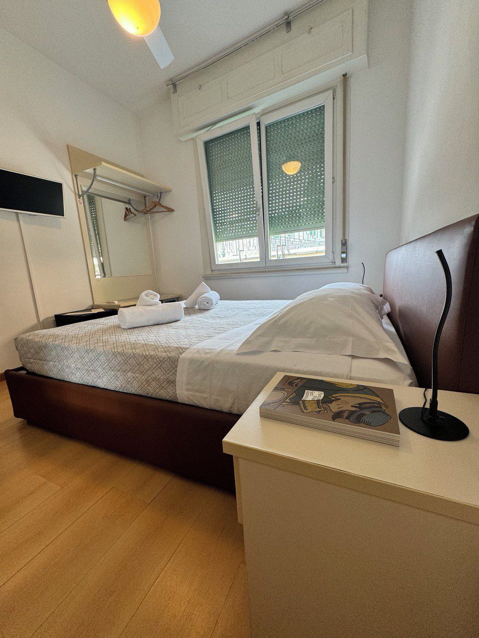 Una habitación de hotel con cama, mesita de noche con un libro, espejo y ventana con persianas.