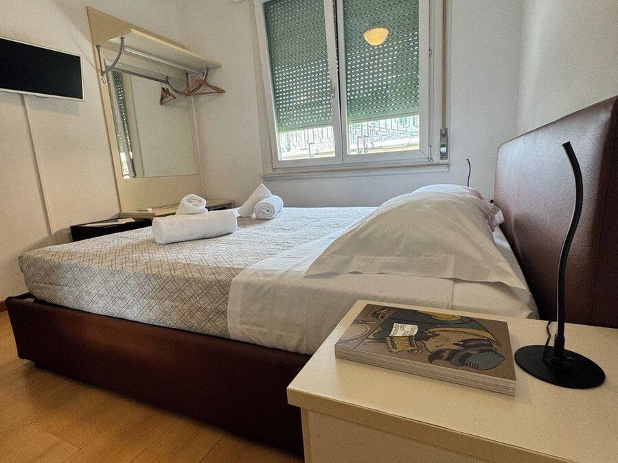 Una habitación de hotel con cama, mesita de noche con un libro, espejo y ventana con persianas.