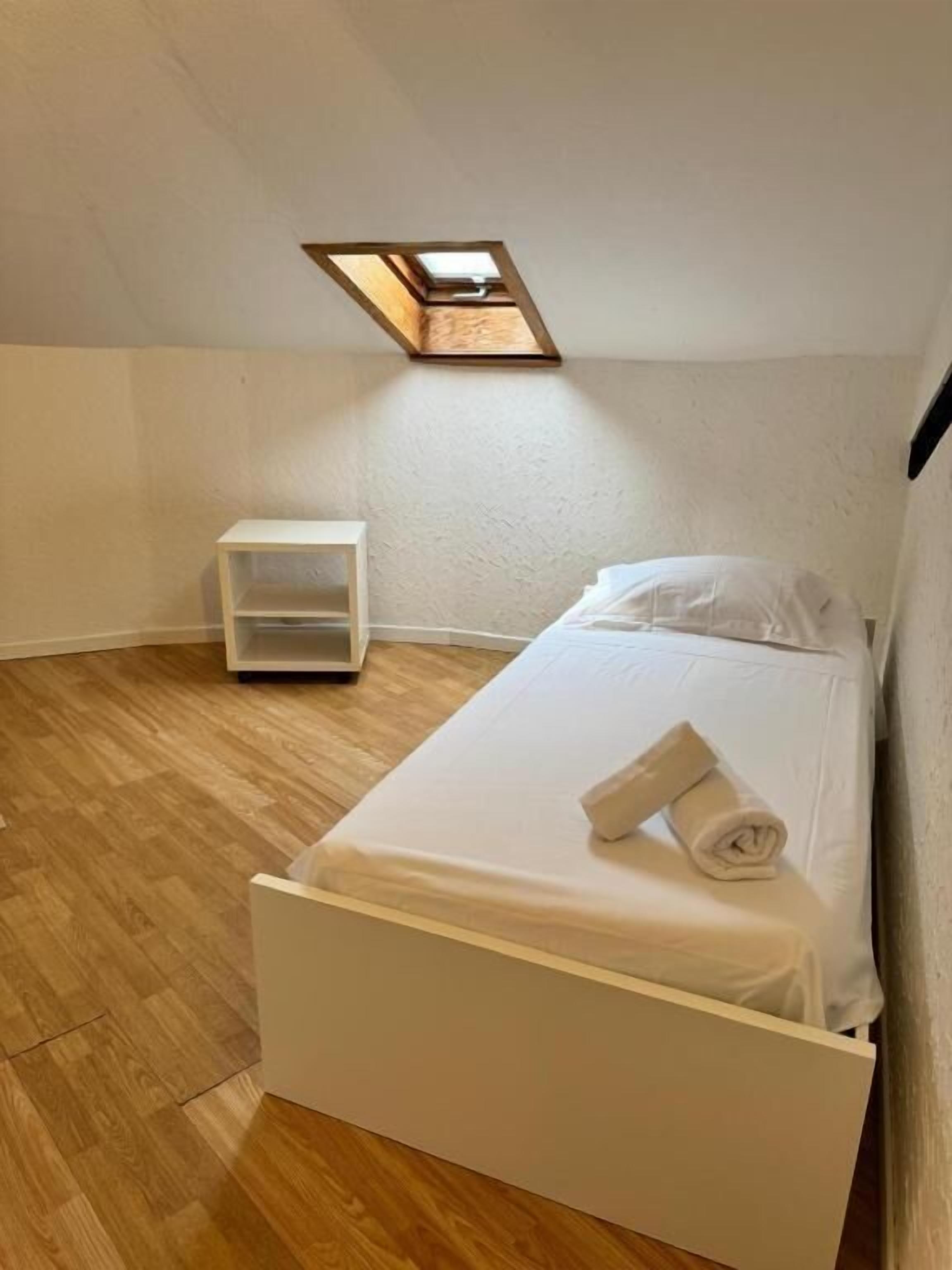 A small, neatly arranged bedroom with a single bed, a nightstand, and a skylight above.