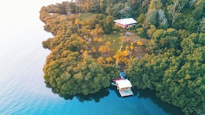 Vista aérea de una isla tropical con densa vegetación, un pequeño bote atracado cerca de un edificio con techo rojo y un mar azul y transparente.