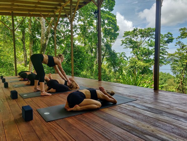 Una terraza de madera con tapetes y bloques de yoga, rodeada de vegetación exuberante y vista al mar.