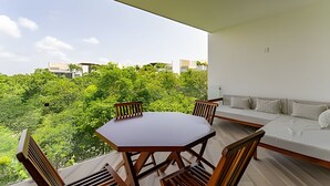 A balcony with a wooden table and chairs, a white sofa, and a view of lush greenery and buildings in the background. - oasis yaaxlum (Tulum)