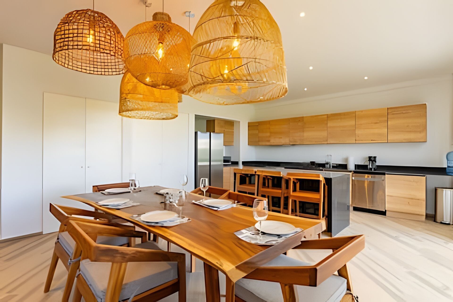 Un espace de salle à manger moderne avec une table et des chaises en bois, une cuisine en arrière-plan et des luminaires suspendus.
