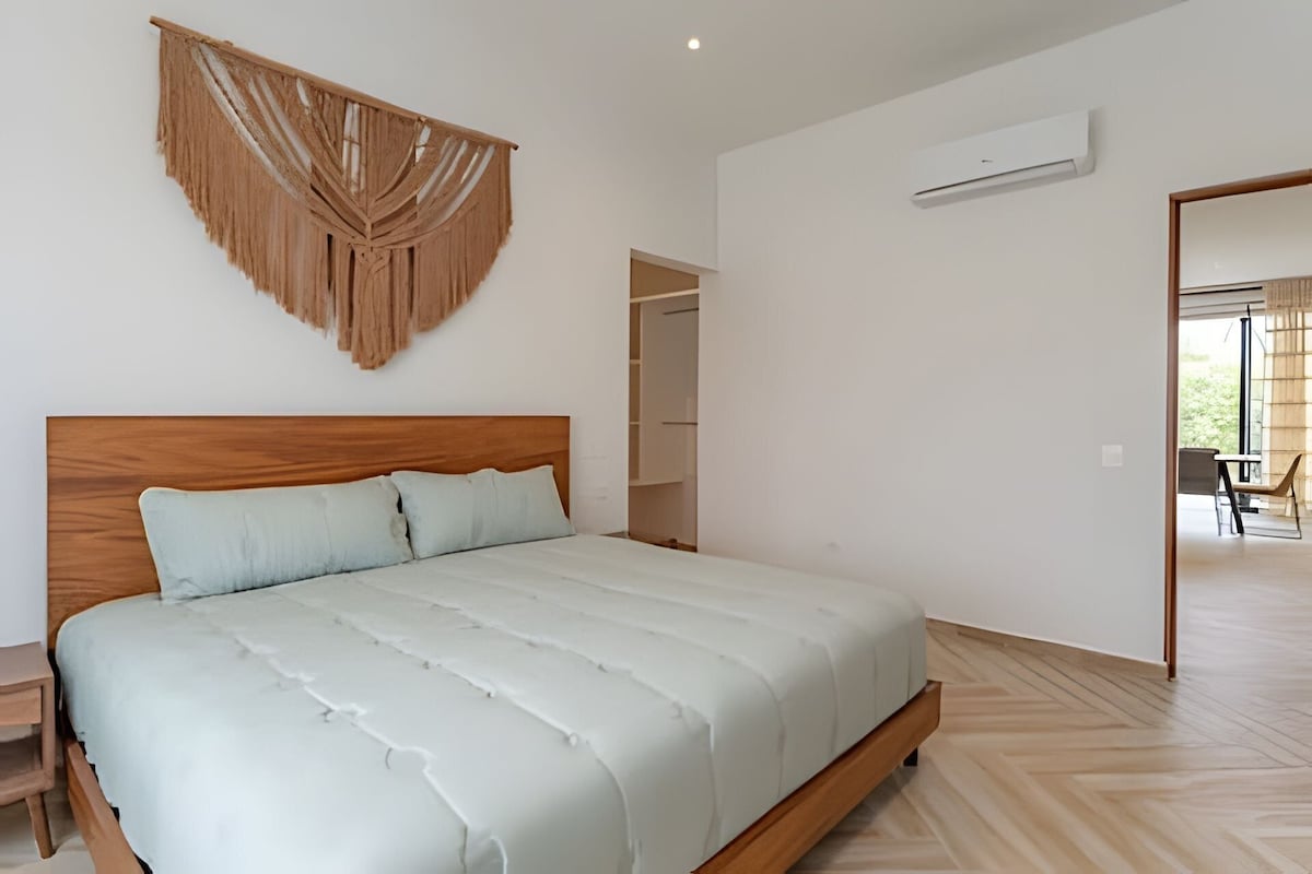 A bedroom with a wooden headboard, a bed with white bedding, a wall-mounted air conditioner, and a view of an outdoor area through an open door.