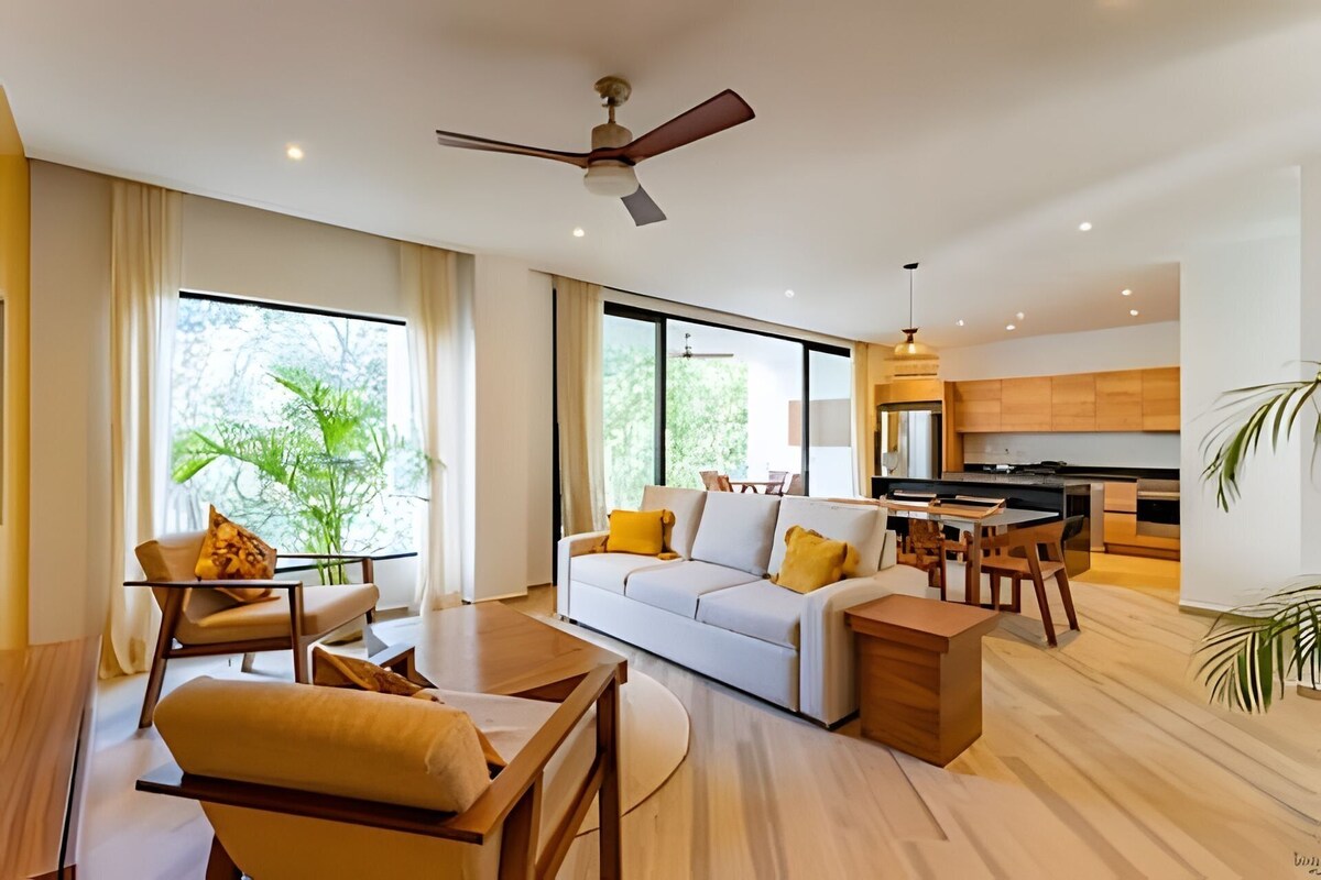 Una sala de estar moderna con sofá, mesa de centro y comedor. Se observa una cocina con armarios de madera y un ventanal grande.
