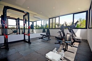 Ett gym med en knäböjsställning, en bänkpress och olika styrketräningsmaskiner, med stora fönster som ger utsikt över palmer.