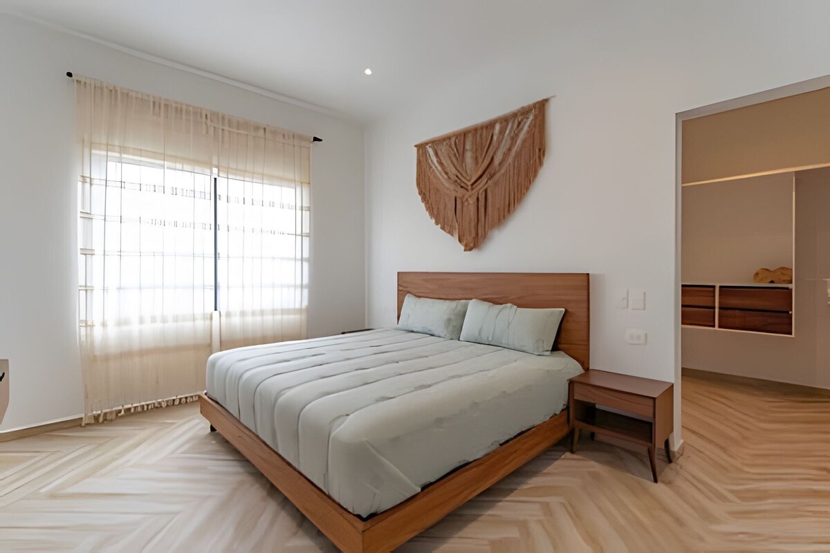 A bedroom with a wooden bed, a nightstand, a large window with sheer curtains, and a wall-mounted decorative piece.