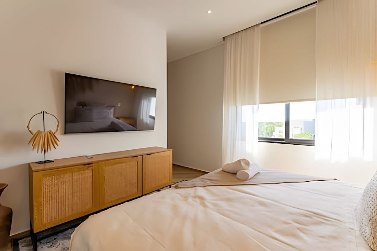 A modern bedroom with a large bed, a flat-screen TV mounted on the wall, a wooden cabinet, and a window with curtains.