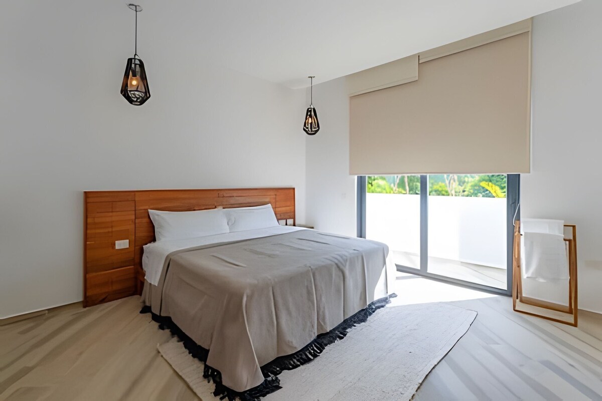 A modern bedroom with a wooden headboard, a large bed with a grey bedspread, two pendant lights, a large window with a view of greenery, and a white bedside table.