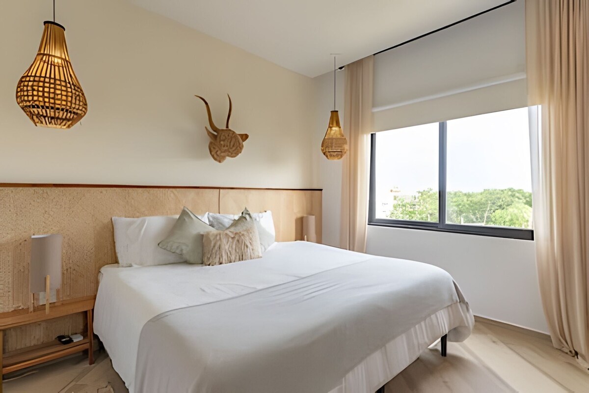 A modern hotel room with a large bed, two wall-mounted lamps, a decorative animal head, and a large window offering a view of greenery.