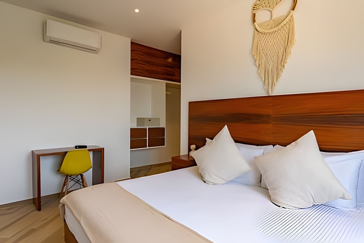 A hotel room with a wooden headboard, a bed with white linens, a yellow chair, and a macrame wall hanging.