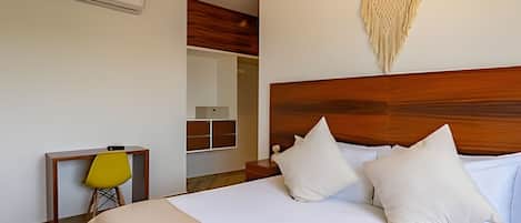 Una habitación de hotel con cabecera de madera, cama con ropa blanca, una silla amarilla y un tapiz de macramé en la pared.