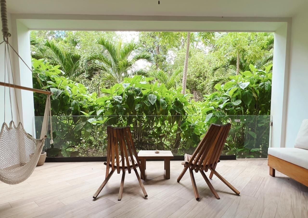 Een kamer met een groot raam, twee houten stoelen, een klein tafeltje en een hangstoel.