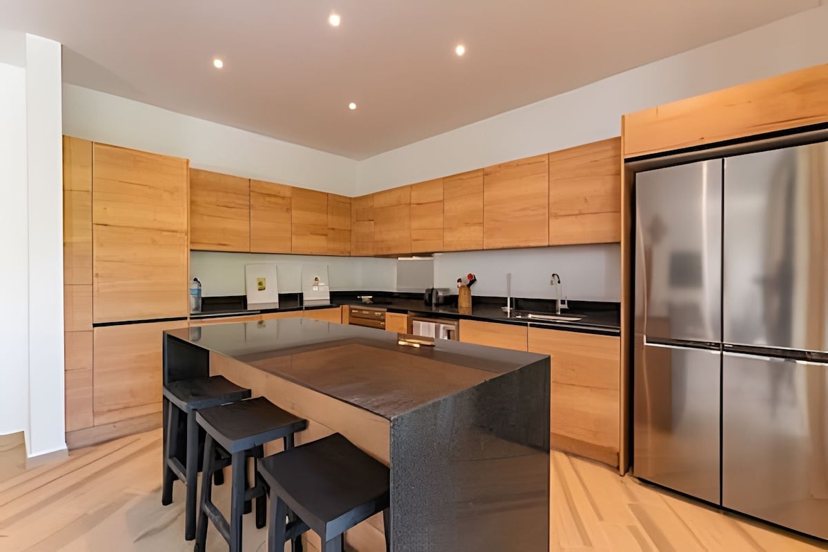 A modern kitchen with wooden cabinets, a central island with stools, a stainless steel refrigerator, and a black countertop.