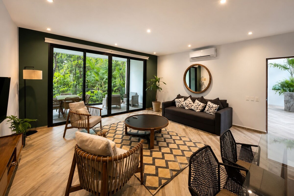 A modern living room with a sofa, chairs, a coffee table, and a view through large sliding doors to a balcony.