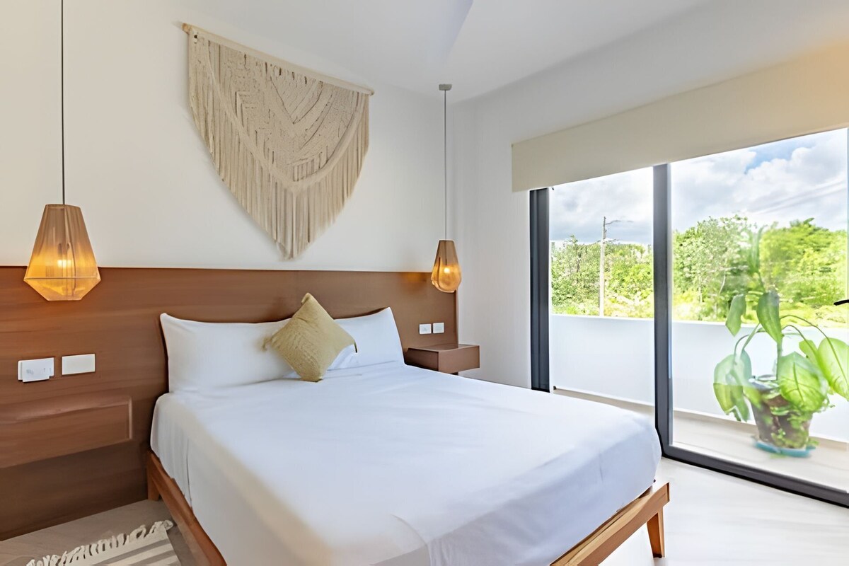 A modern bedroom with a large bed, wooden headboard, two bedside lamps, and a view of greenery through large windows.