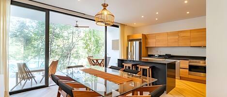 A modern kitchen with wooden cabinets, a glass dining table, and a large window overlooking a forest.