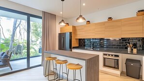 A modern kitchen with wooden cabinets, a central island with stools, a black backsplash, and a large window offering a view of greenery. - oasis yaaxlum (Tulum)