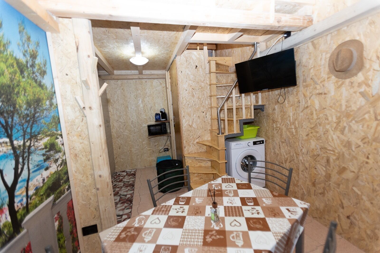 Ein gemütlicher Raum mit Holzboden, einem Tisch mit kariertem Tuch, einer Waschmaschine, einer Mikrowelle und einem an der Wand befestigten Fernseher.