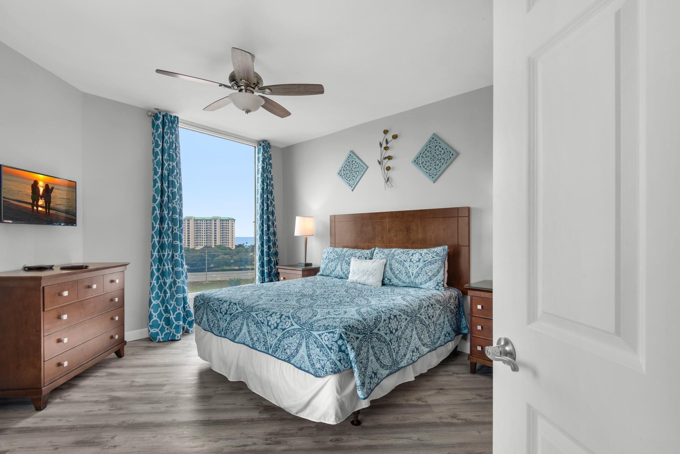 A bedroom with a large bed, wooden headboard, a ceiling fan, a nightstand with a lamp, a dresser, and a window with curtains.