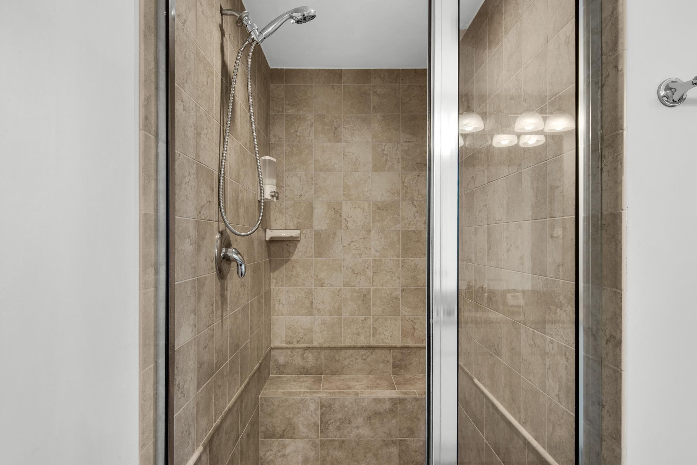A modern shower with a glass door, beige tiles, and a handheld showerhead.