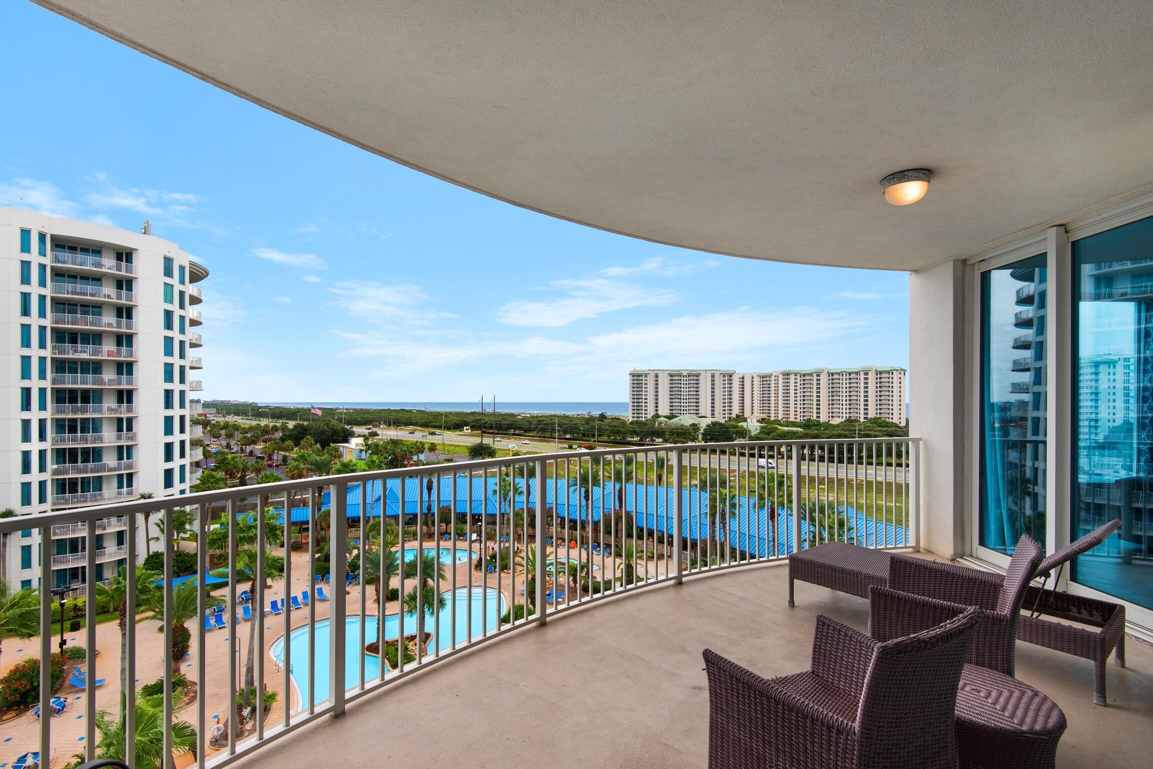 A balcony with wicker furniture, a pool, and a view of a resort area with buildings.