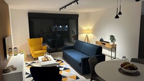 A modern living room with a sofa, a yellow armchair, a dining table with a fruit bowl, and a lamp.