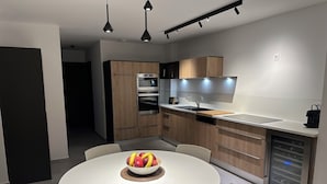 A modern kitchen with wooden cabinets, a built-in oven, and a white dining table with a bowl of fruit.