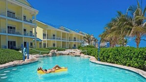 Una piscina con una persona recostada en un flotador amarillo, rodeada de jardines con palmeras y un edificio con balcones al fondo.