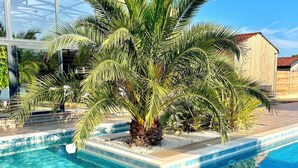 A swimming pool with clear blue water, surrounded by a tiled deck and palm trees. - NUIKI Lodges (Montsegur-sur-Lauzon)