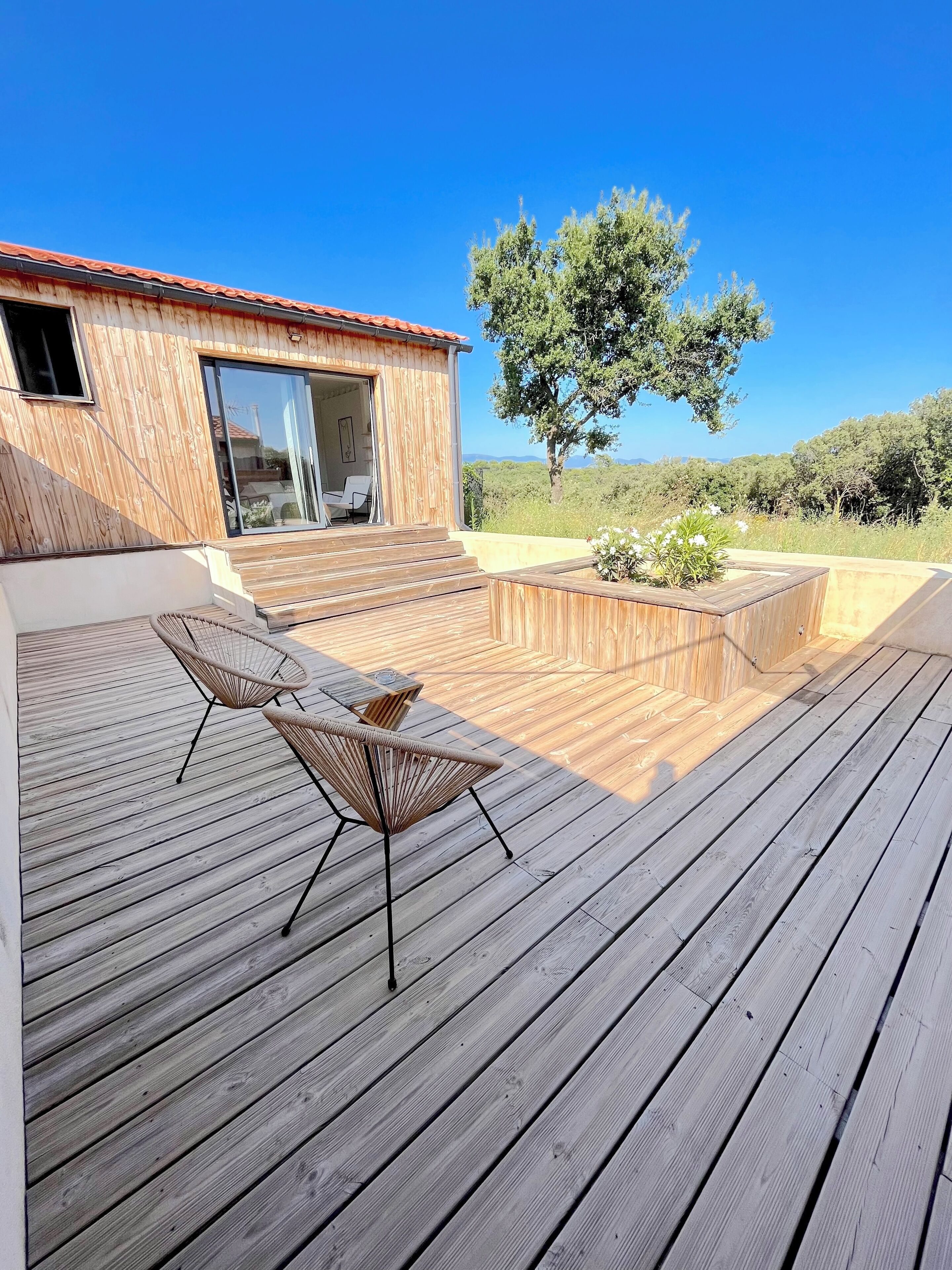 En träveranda med två flätade fåtöljer och ett litet bord, en träbyggnad med ett rött tak och ett stort träd i bakgrunden.