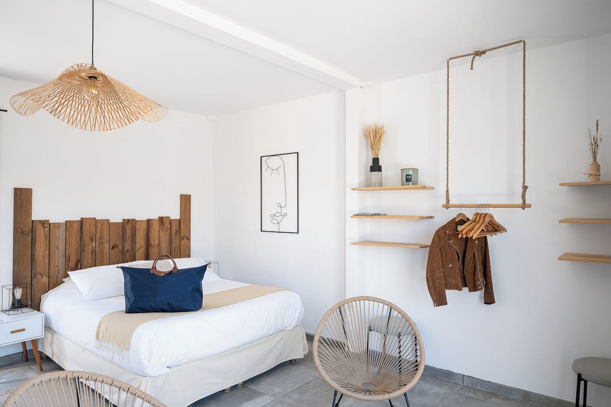 A modern bedroom with a wooden headboard, a bed with white bedding and a blue pillow, a wicker chair, and a hanging light fixture.