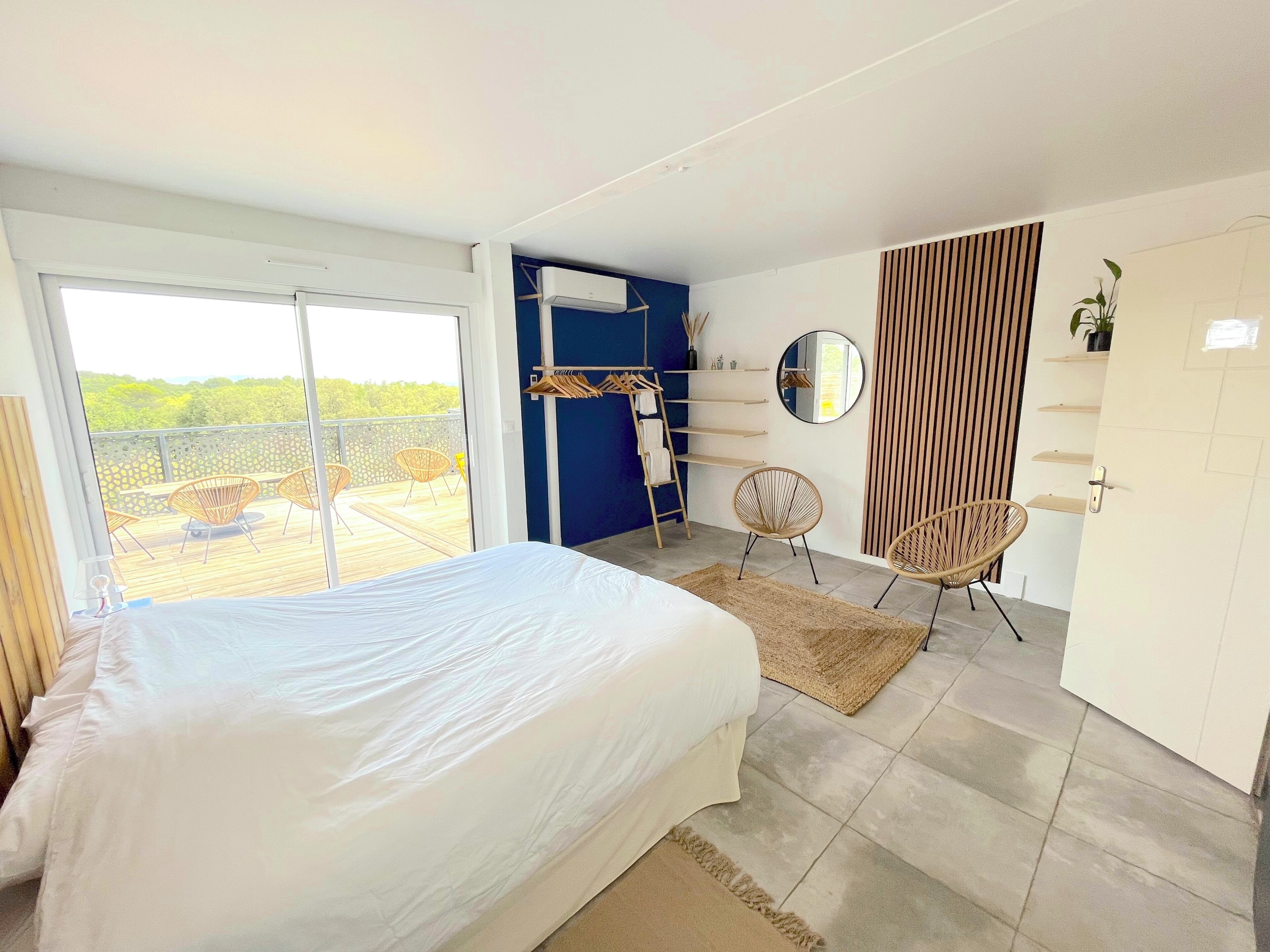 A bedroom with a large bed, a balcony with chairs, a wardrobe, and a mirror.