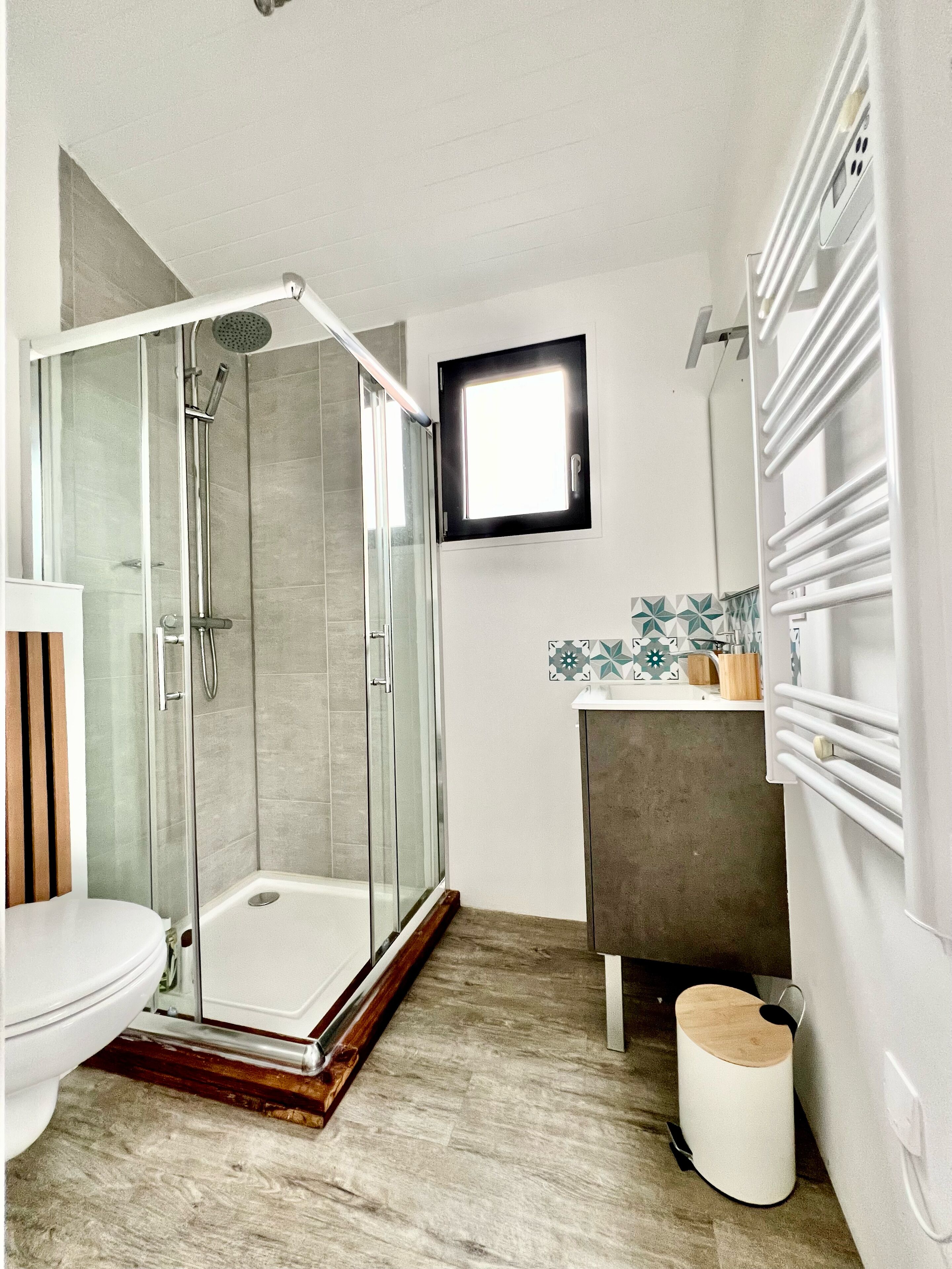 A modern bathroom with a glass-enclosed shower, a toilet, a sink with a tiled backsplash, and a small window.