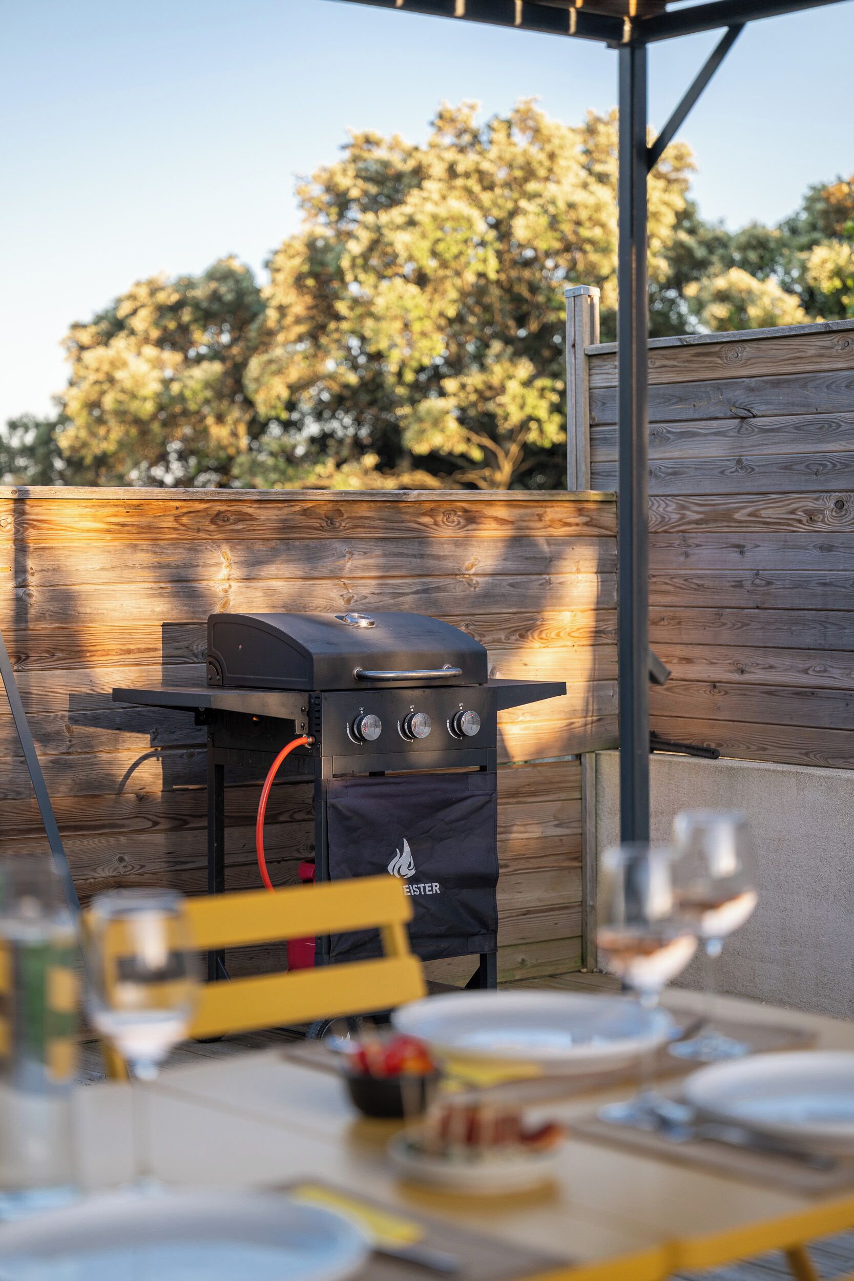 Un espace extérieur aménagé pour les repas, avec un barbecue, une clôture en bois et une table dressée pour deux personnes.