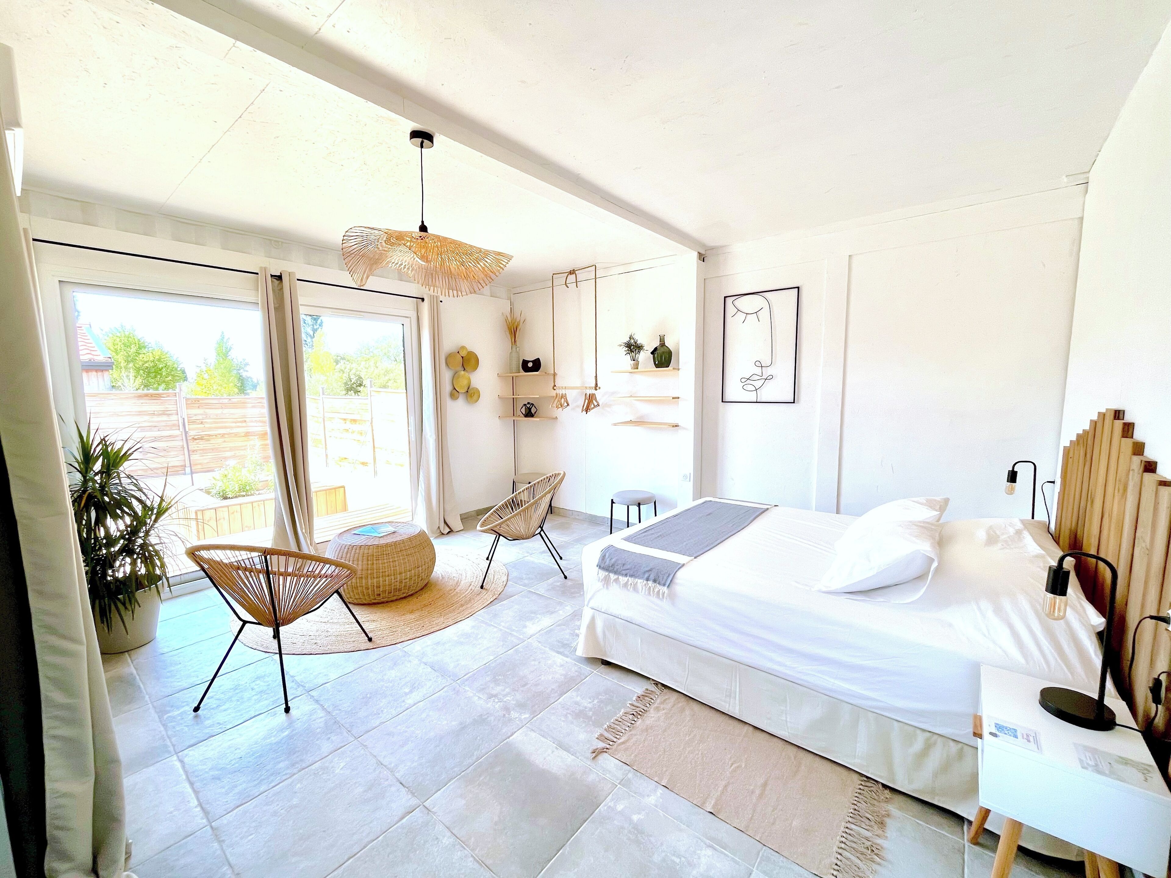 A bright, modern bedroom with a large bed, a chair, a small table, and a view of the outdoors.