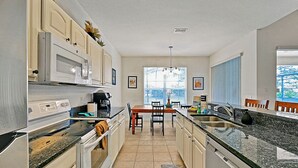Una cocina moderna con armarios blancos, encimera de granito, comedor con mesa y sillas de madera, y un ventanal con persianas.